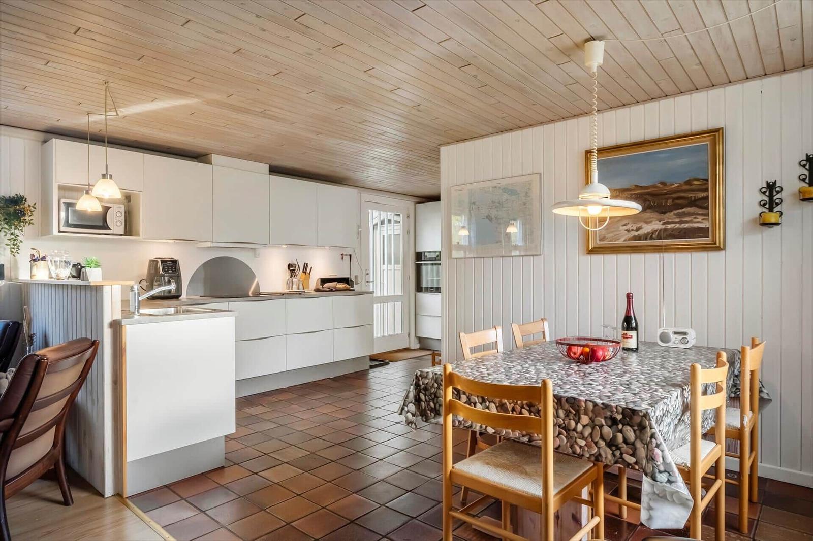 Küche und Essbereich mit Holzdecke, weißen Schränken und Steinbelag am Tisch.