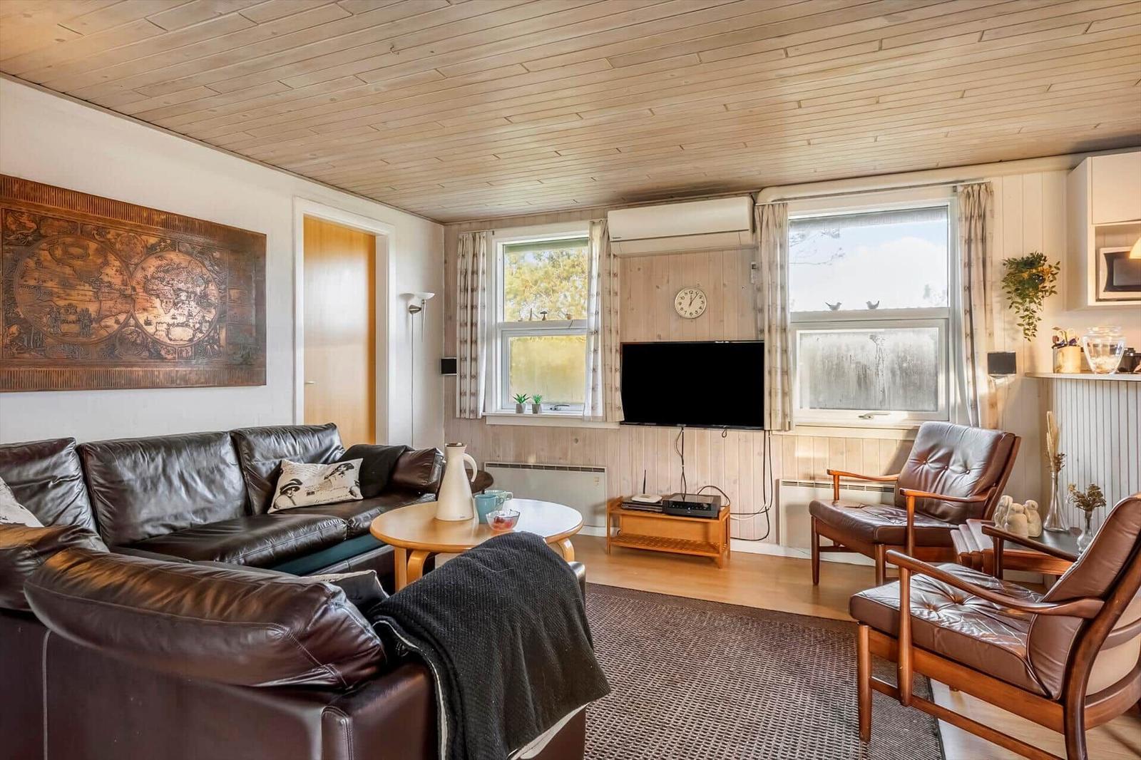 Wohnzimmer mit Ledersofa, zwei Sessel und Fernseher. Holzwände und Decke. Fenster mit Blick draußen.