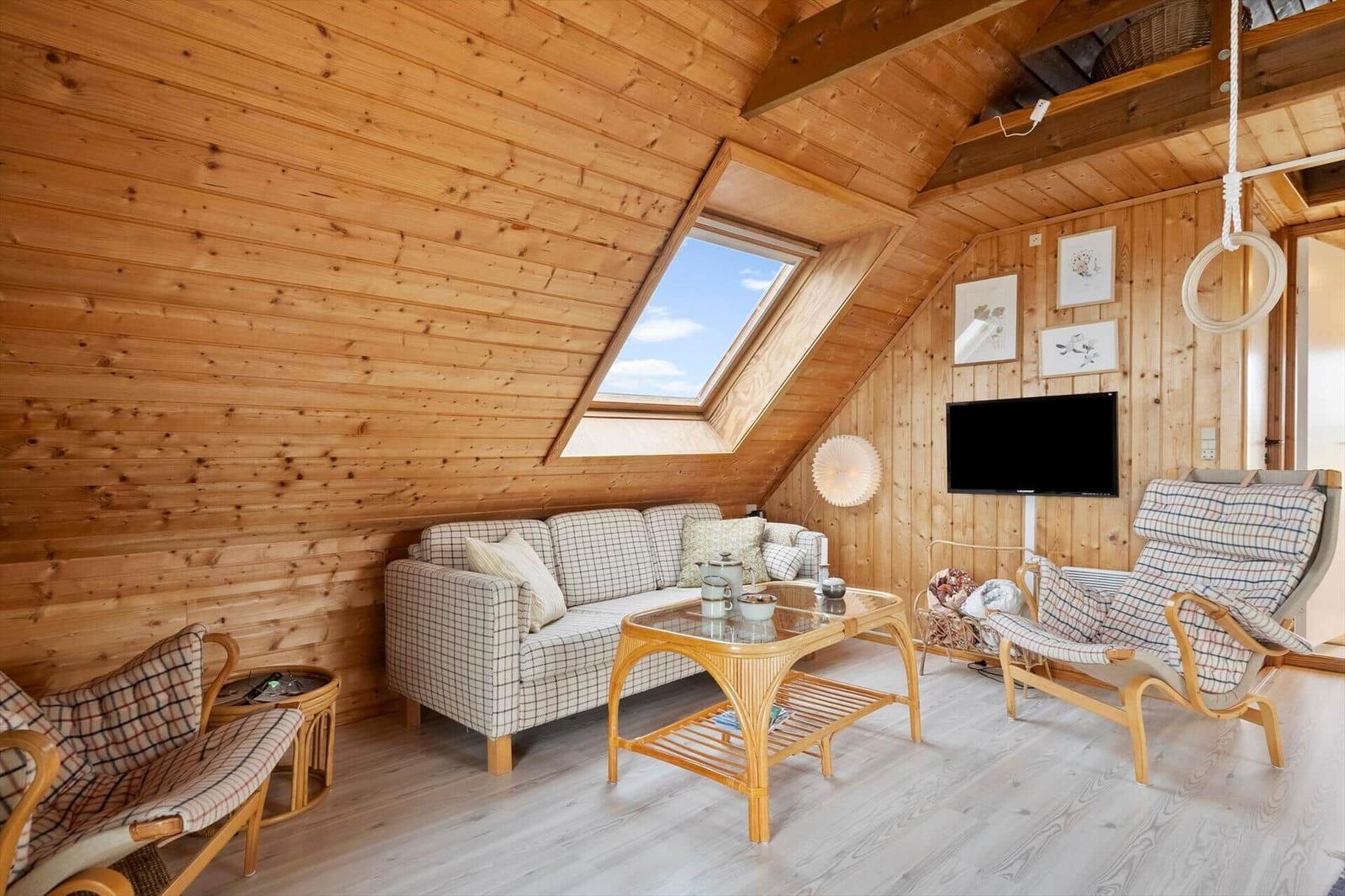 Wohnzimmer mit Holzwand, Sofa, Sessel und TV. Dachfenster und helle Bodenplanken.