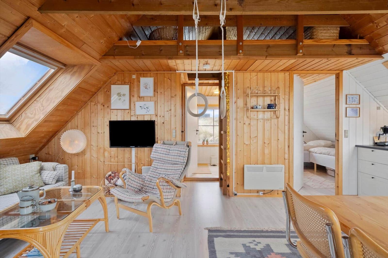 Wohnraum mit Holzwänden, Sofa, Sessel und Fernseher. Durchgang zu Schlafzimmer und Küche.