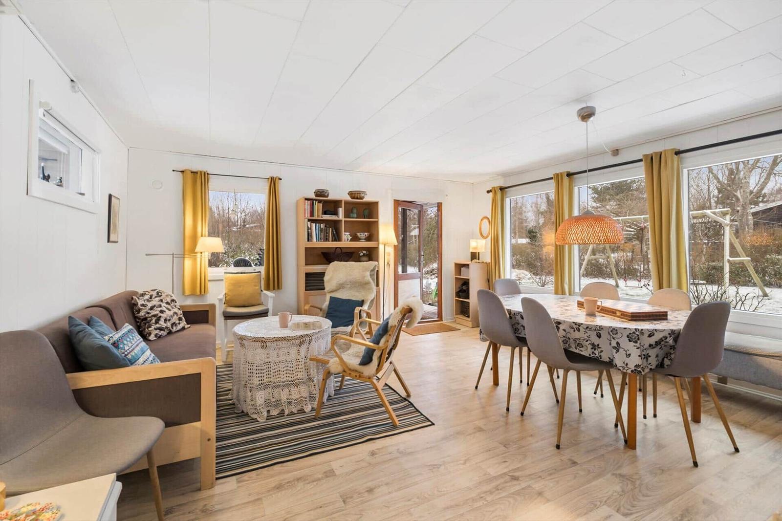 Wohn- und Essbereich mit Sofa, Stühlen und Tisch. Große Fenster mit gelben Vorhängen.