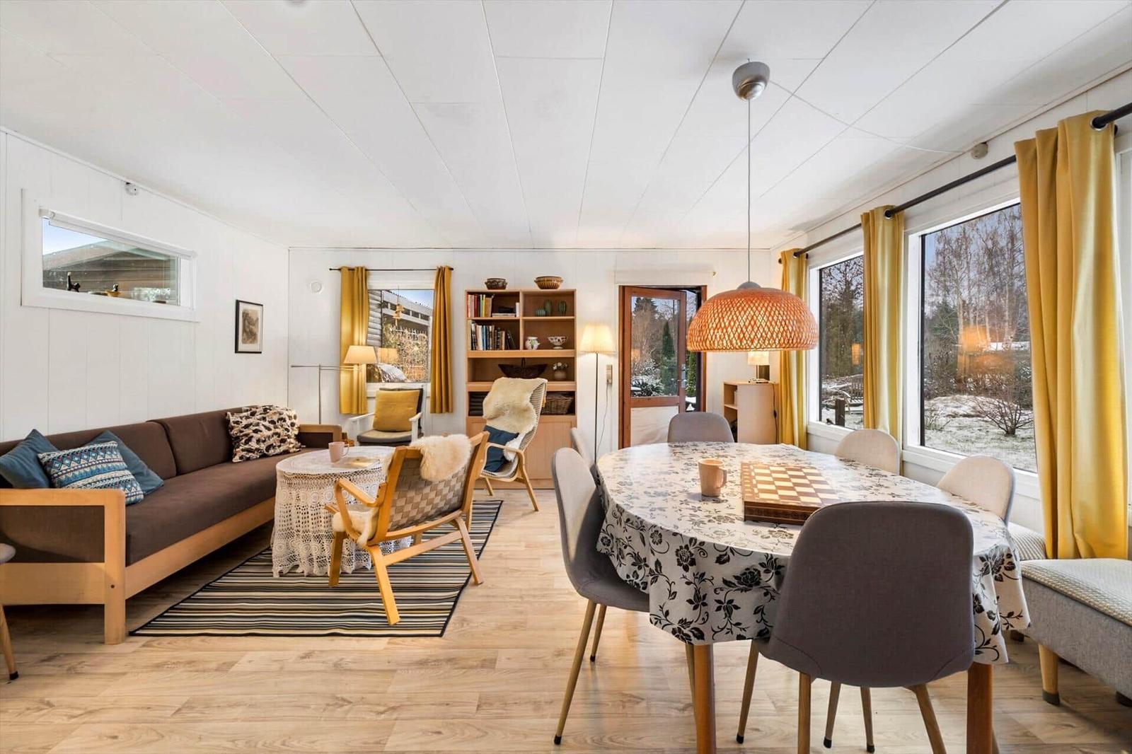 Wohn- und Essbereich mit Sofa, Tisch und Stühlen, großen Fenstern und gelben Vorhängen.
