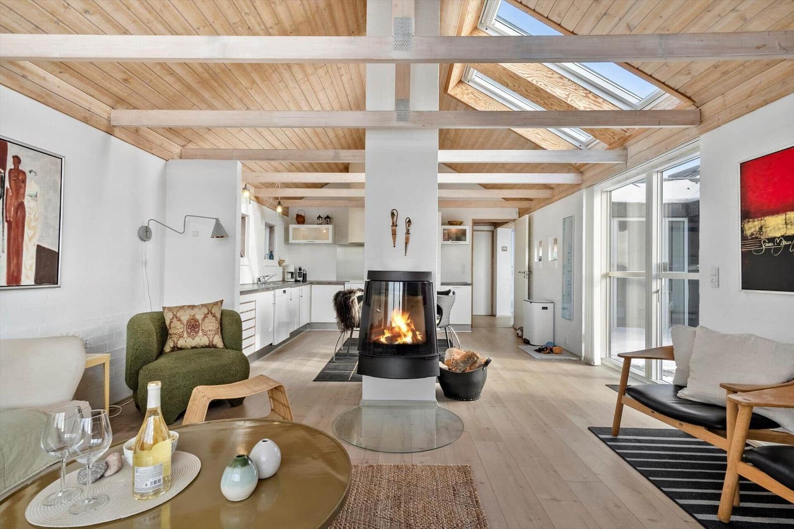 Wohnzimmer mit Kamin, Holzdecke und großem Fenster. Küche im Hintergrund.