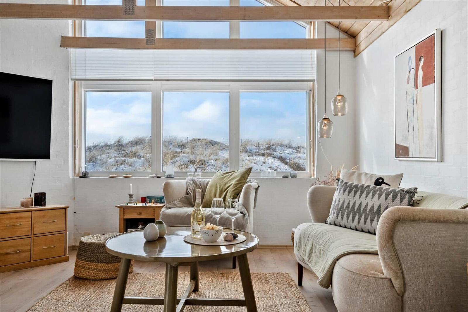 Wohnzimmer mit Sofa, Tisch und Fensterblick auf Dünen.