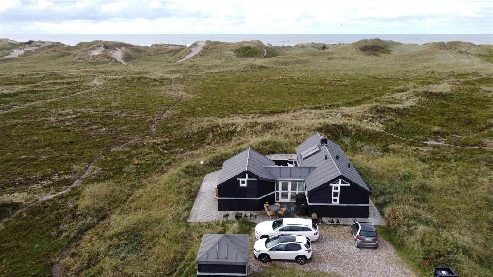 Schwarzes Haus mit Terrasse und Parkplatz in Dünenlandschaft mit Blick aufs Meer.