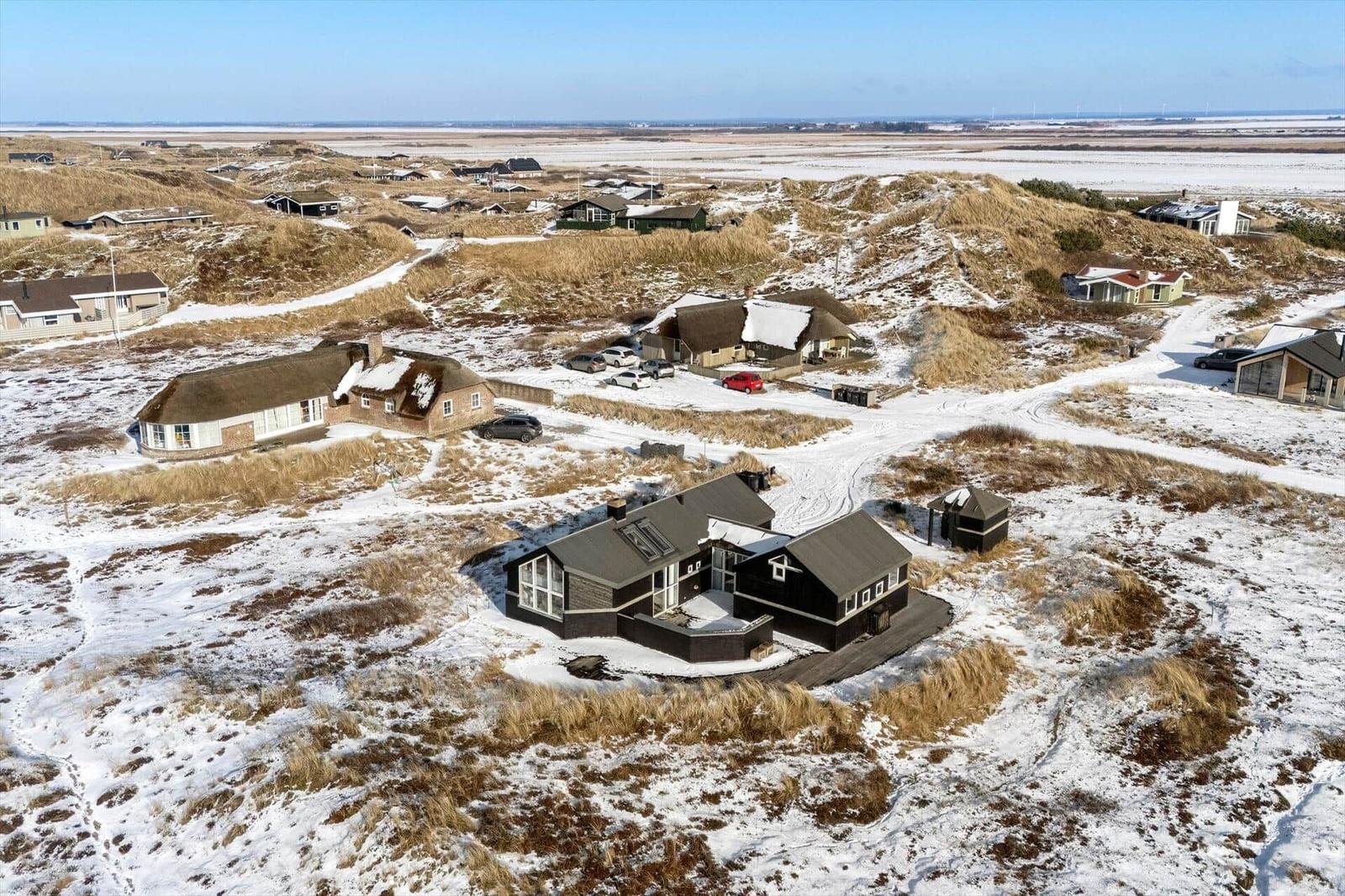 Schneebedeckte Häuser auf Dünenlandschaft mit Blick auf Watt und Windkraftanlagen.