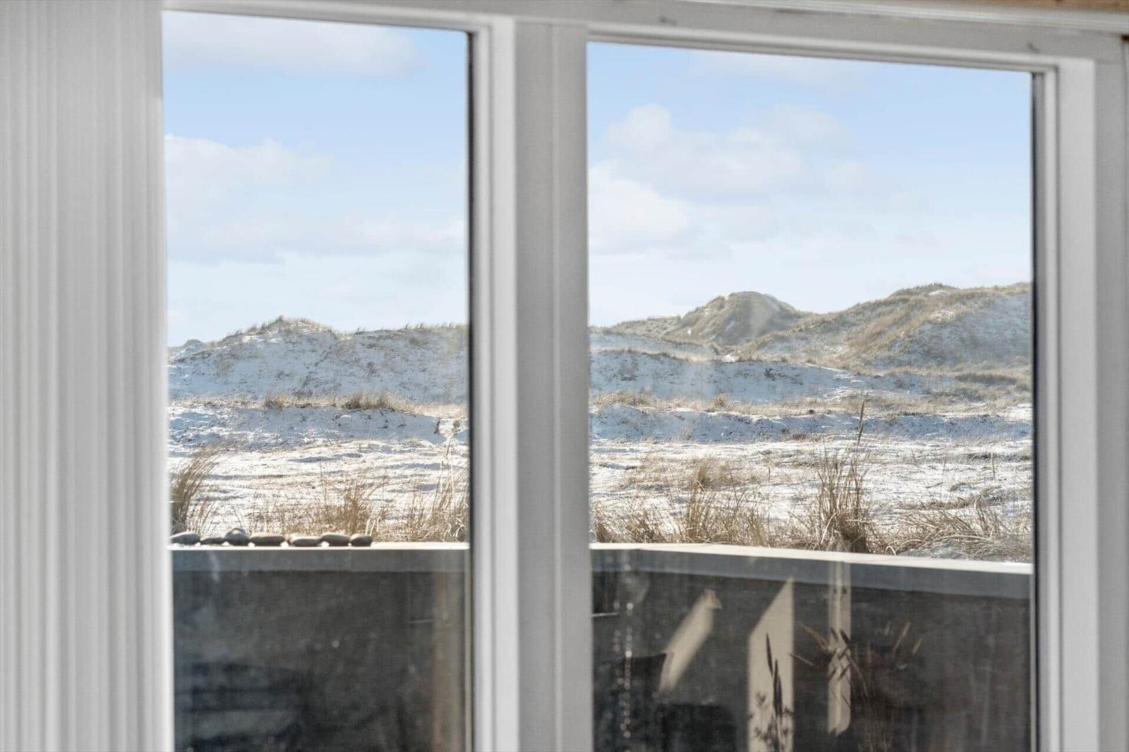 Durch das Fenster blickt man auf sandige Dünen mit trockenem Gras.
