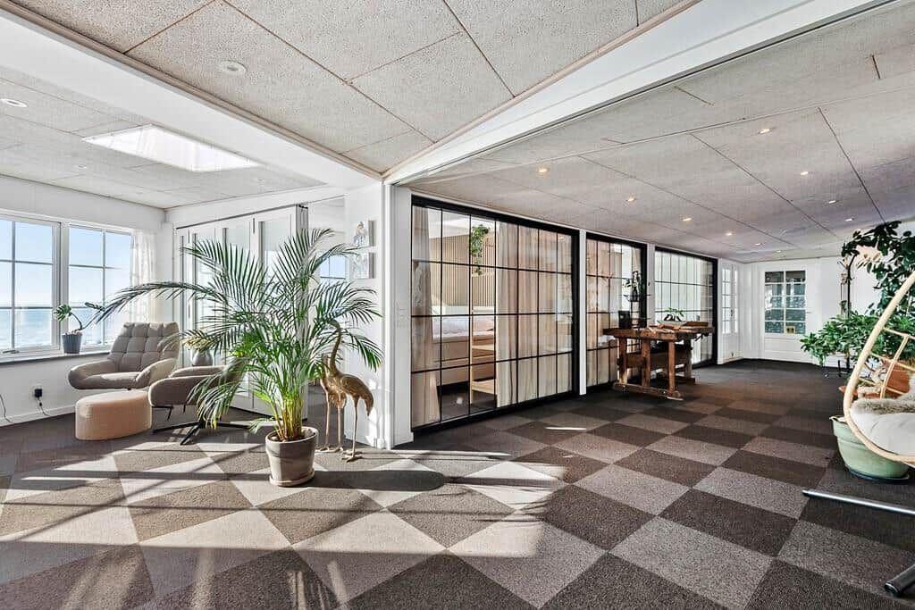Large room with glass partition, seating, and plants. Checkered floor and windows with sea view.