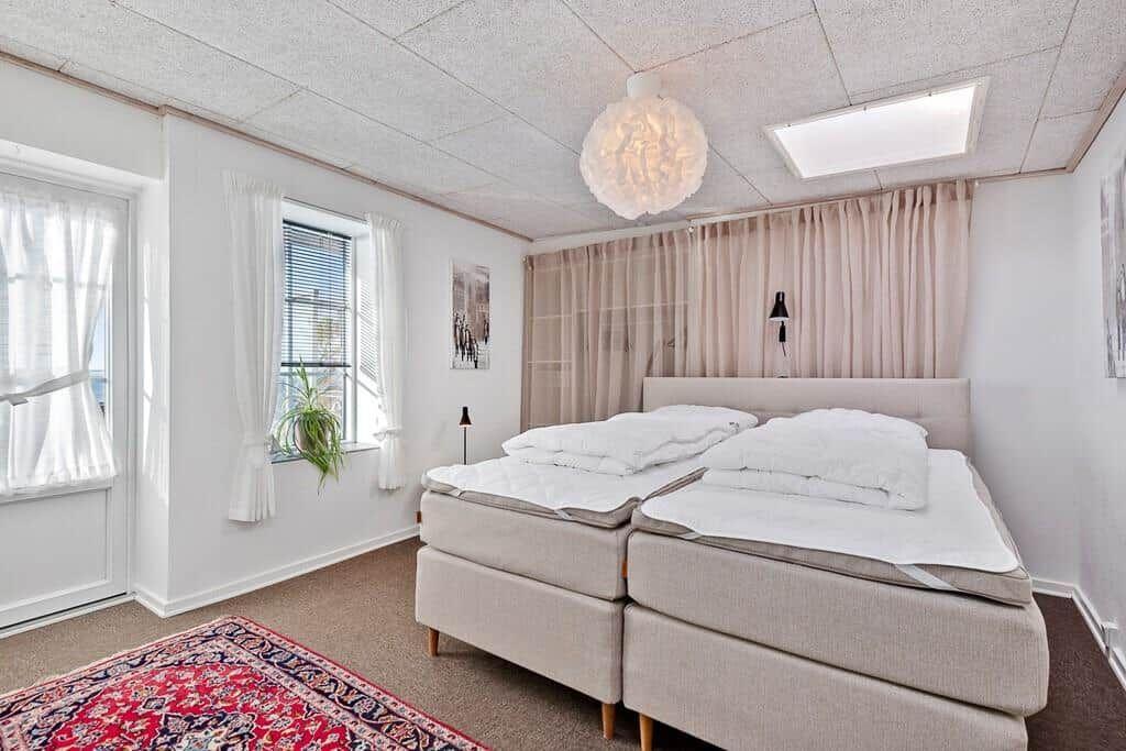Two beds with white bedding, windows with curtains, and rug on the floor.