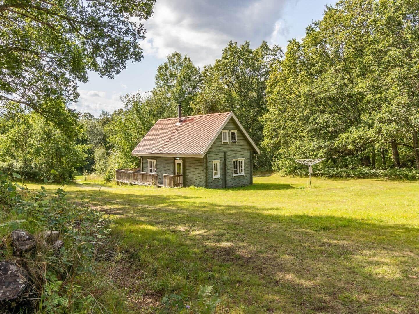 Holzhütte mit Terrasse im Grünen. Umgeben von Bäumen und Grasfläche.