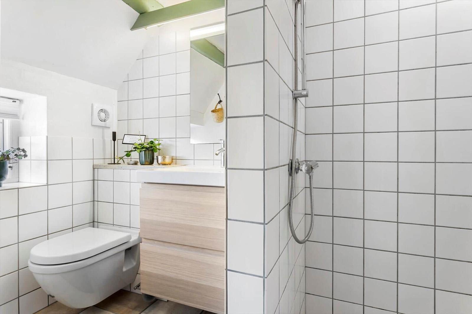Badezimmer mit Dusche, Waschbecken und Toilette. Weiße Fliesen und Holzfronten.