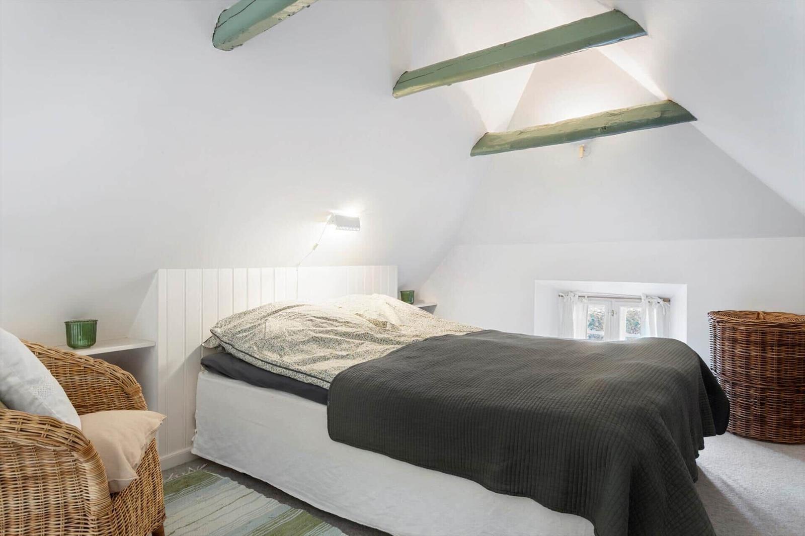 Schlafzimmer mit Bett, Wickerstuhl und grünen Akzenten unter schrägen Deckenbalken.