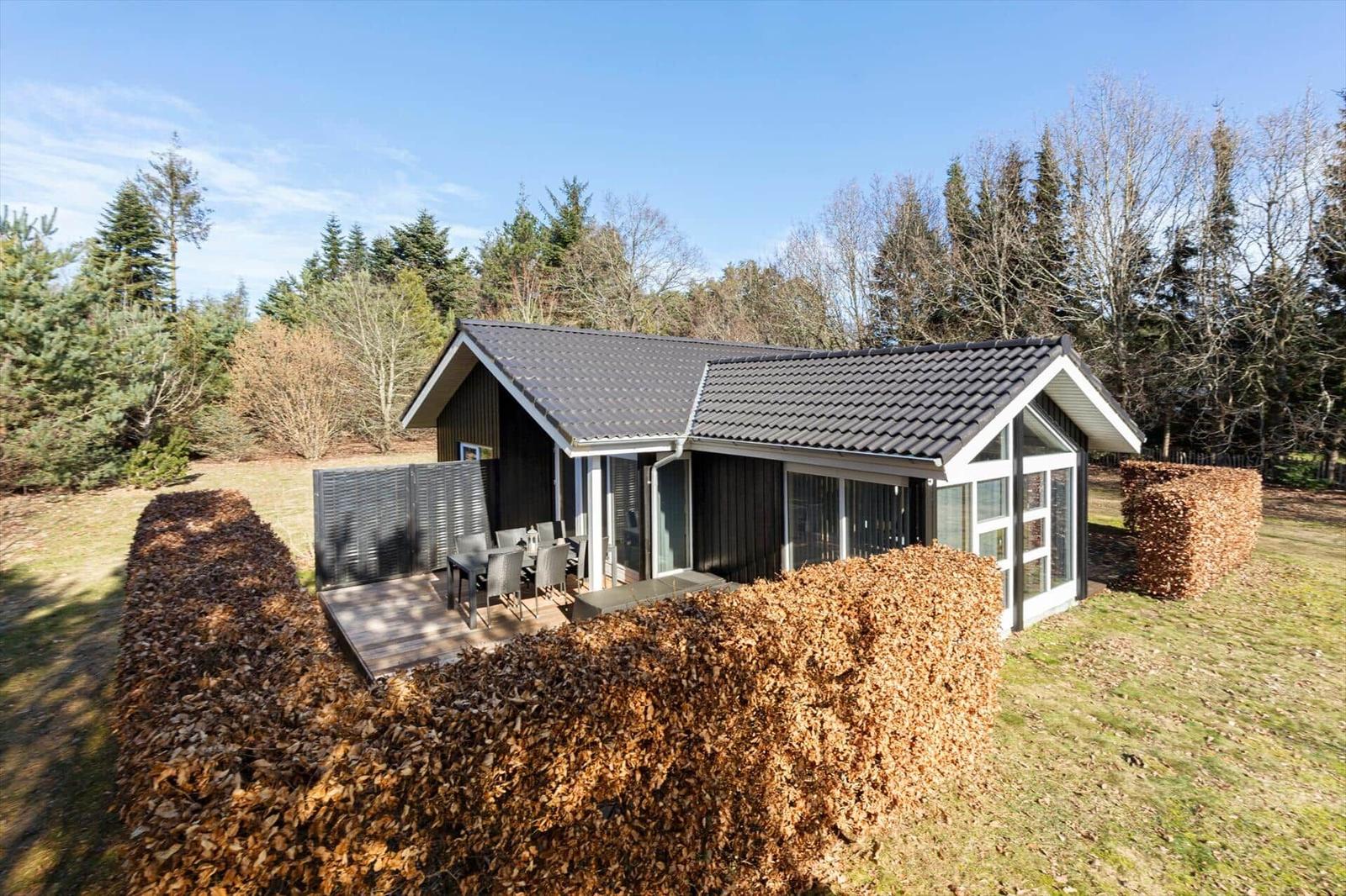 Modern cabin with terrace and seating, surrounded by hedges and trees.