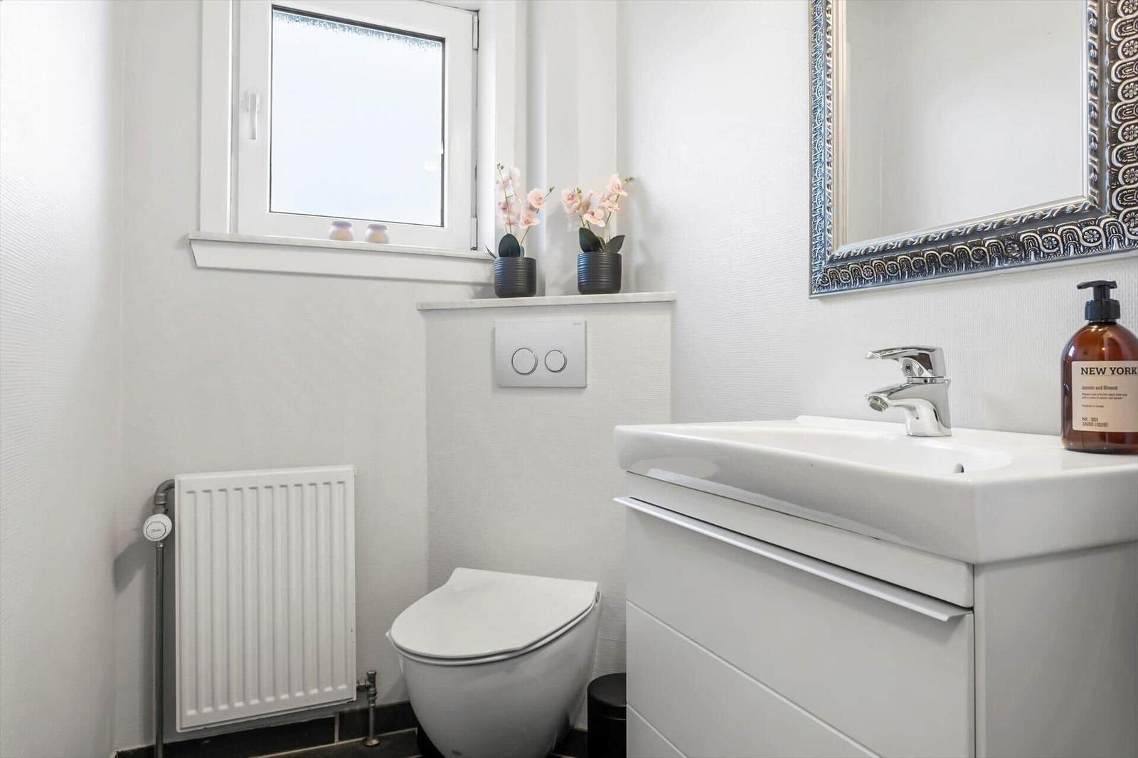 Badezimmer mit Toilette, Waschbecken und Spiegel. Fenster mit Blumen auf Sims.