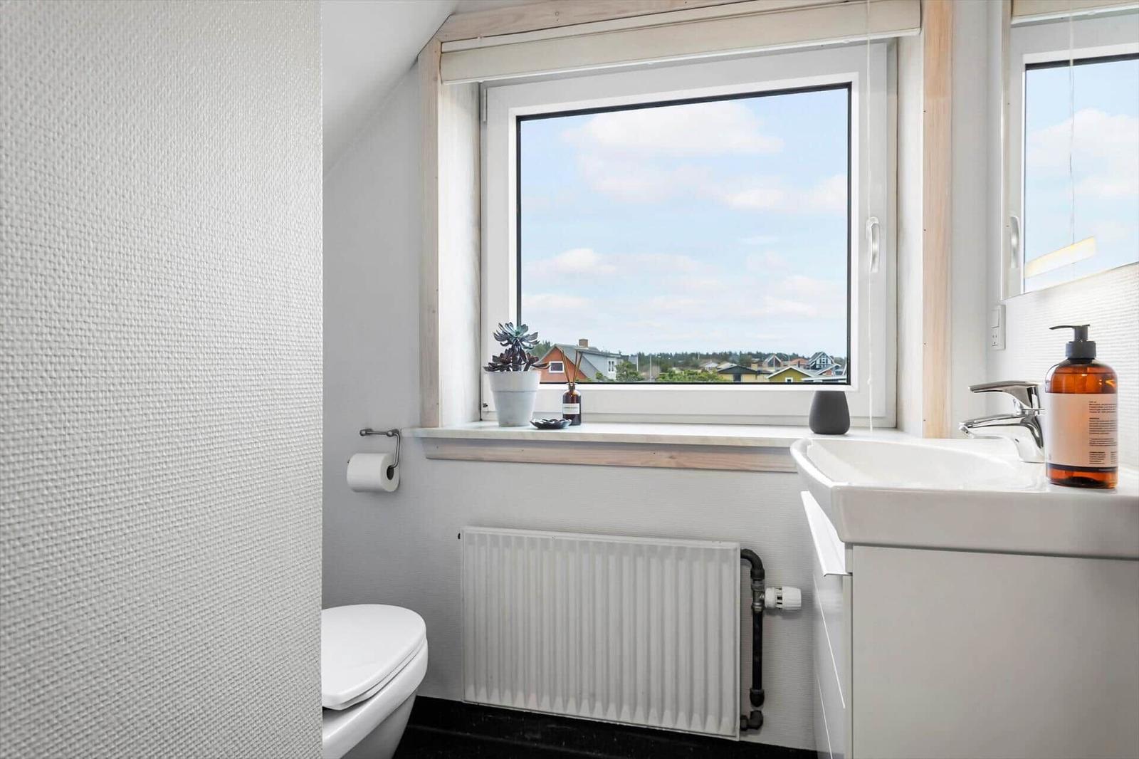 Badezimmer mit Fenster, Waschbecken und Toilette. Draußen sind Häuser und Bäume sichtbar.