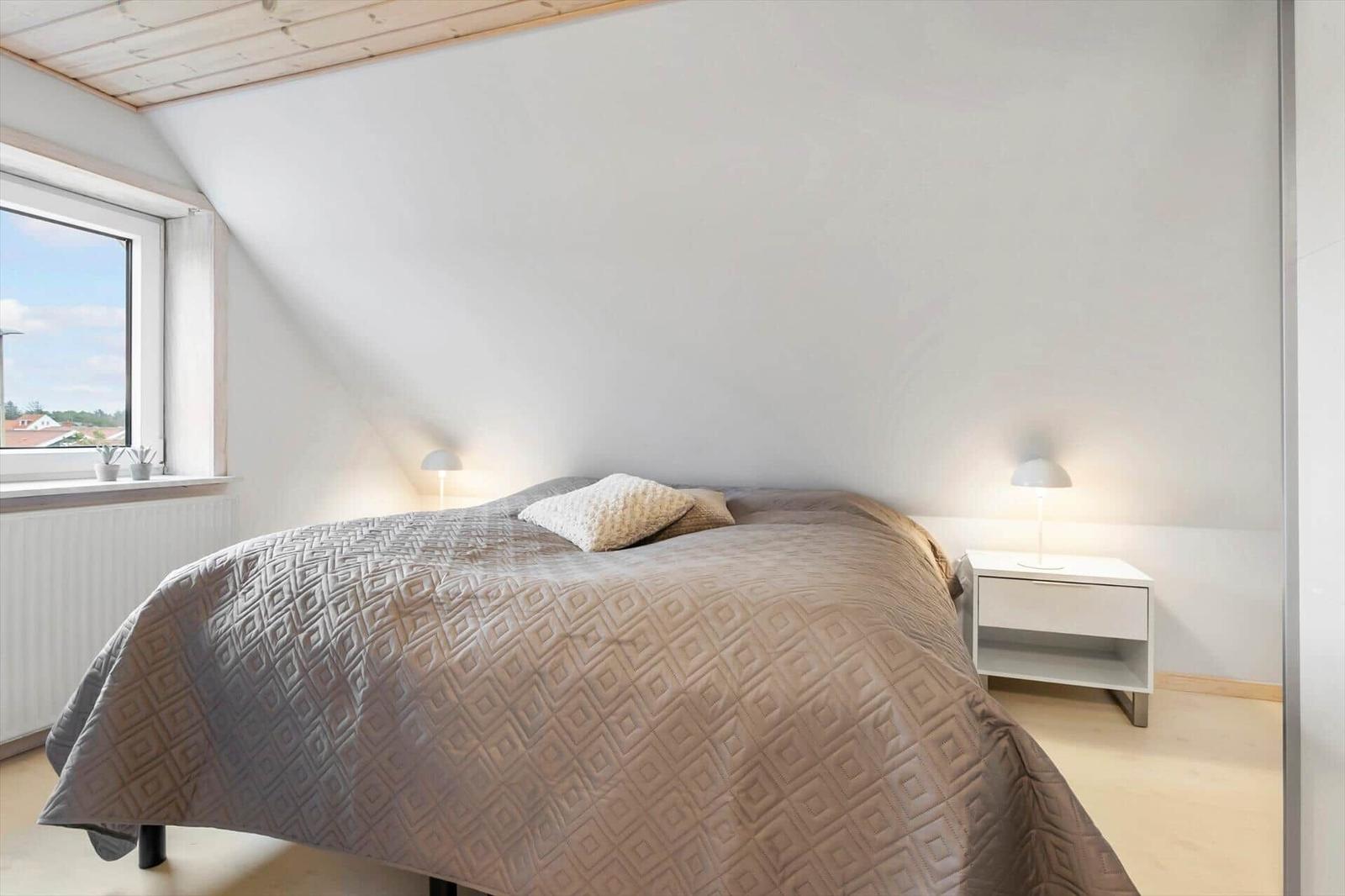 Schlafzimmer mit Bett, Nachttisch und Fenster. Holzdecke und helles Licht.