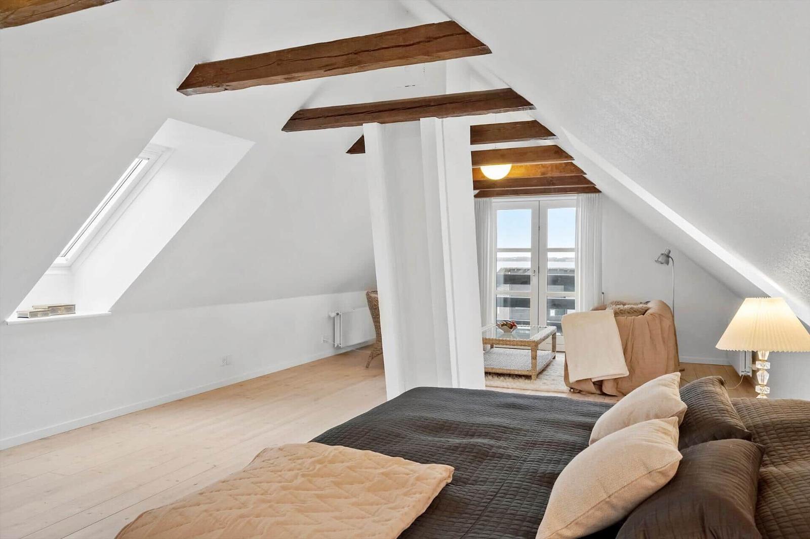 Schlafzimmer mit Dachschrägen, Holzbalken und Fenster. Bett, Sofa und Lampen sichtbar.