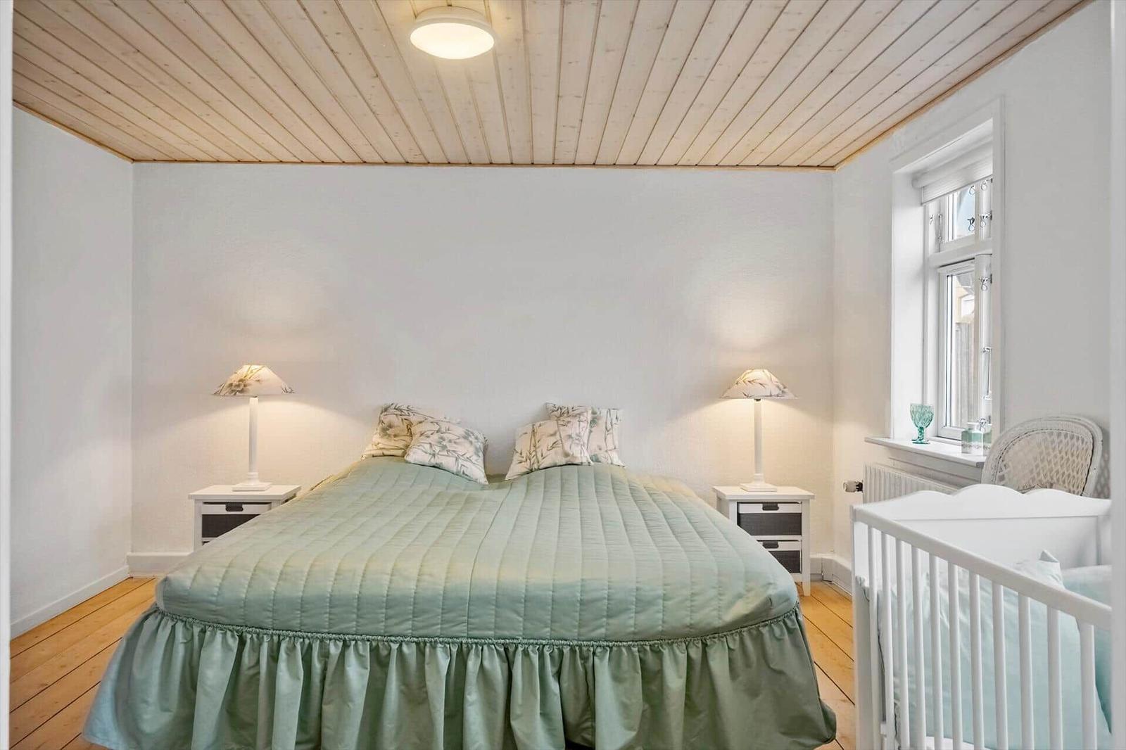 Schlafzimmer mit Bett, Nachttischen und Babybett. Holzdecke und Fenster sichtbar.