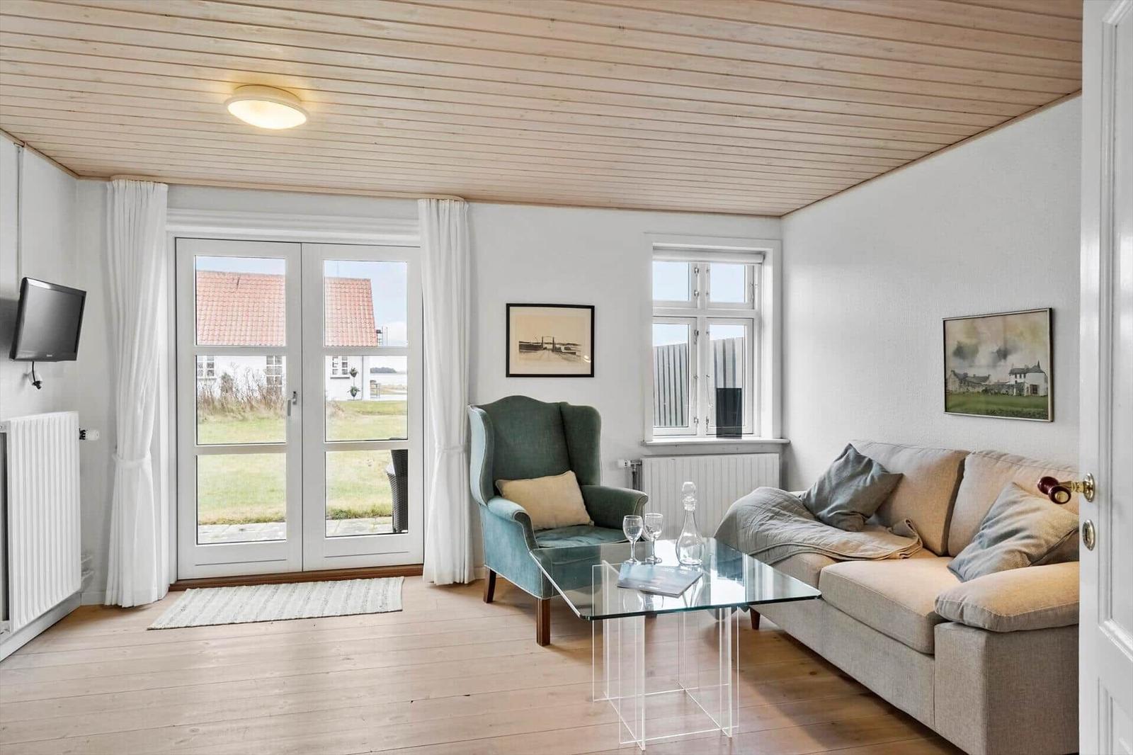 Wohnzimmer mit Sofa, Sessel und Glas-Tisch. Holzdecke und große Fenster.