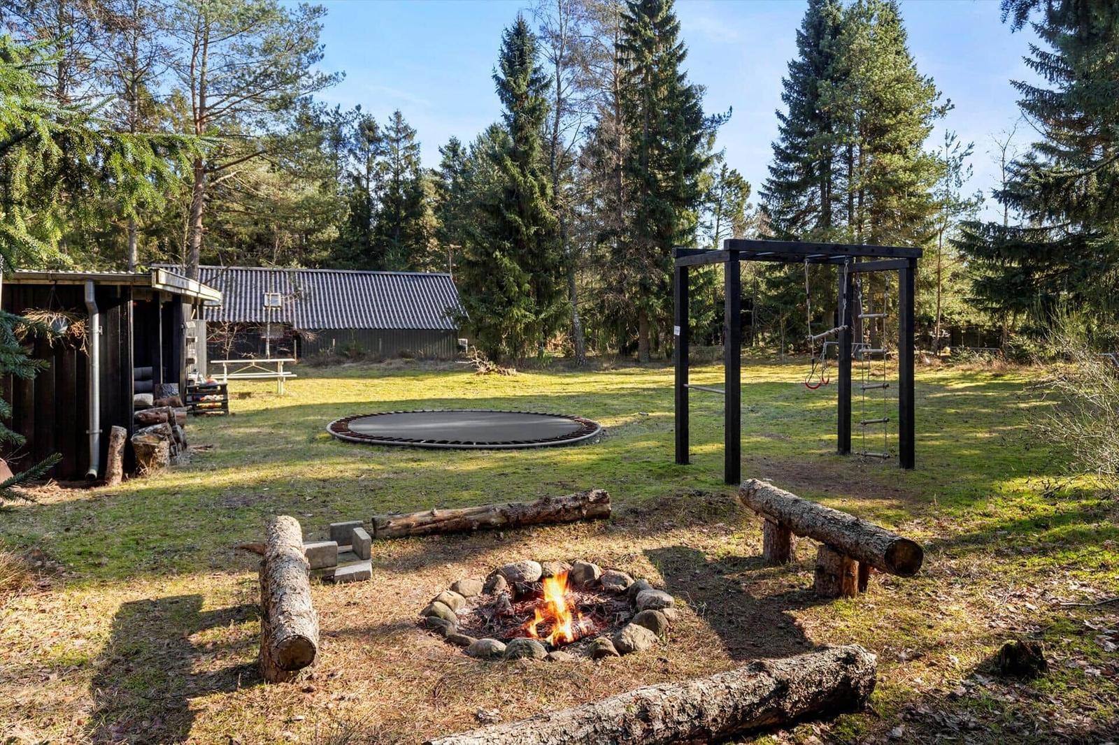 Fire pit, trampoline, and playground in the garden with log bench and trees.
