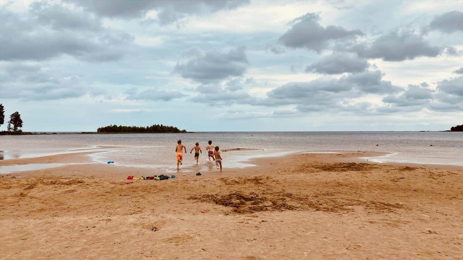 Vier Kinder laufen am Strand entlang, im Hintergrund See und bewölkter Himmel.