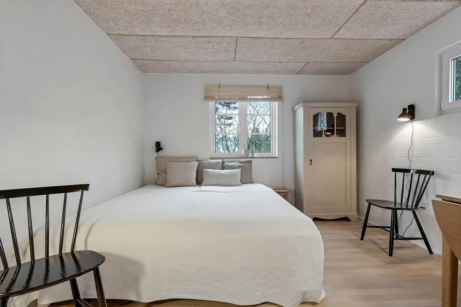 Schlafzimmer mit Bett, Schrank und zwei Stühlen. Fenster mit Blick auf Bäume.