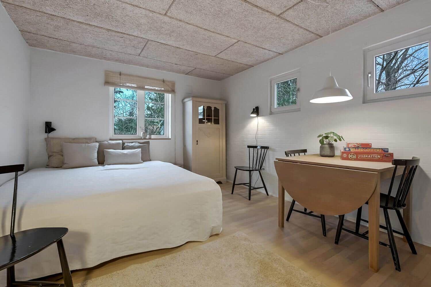 Schlafzimmer mit Bett, Tisch, Stühlen und Fenstern. Decke aus Holzplatten.