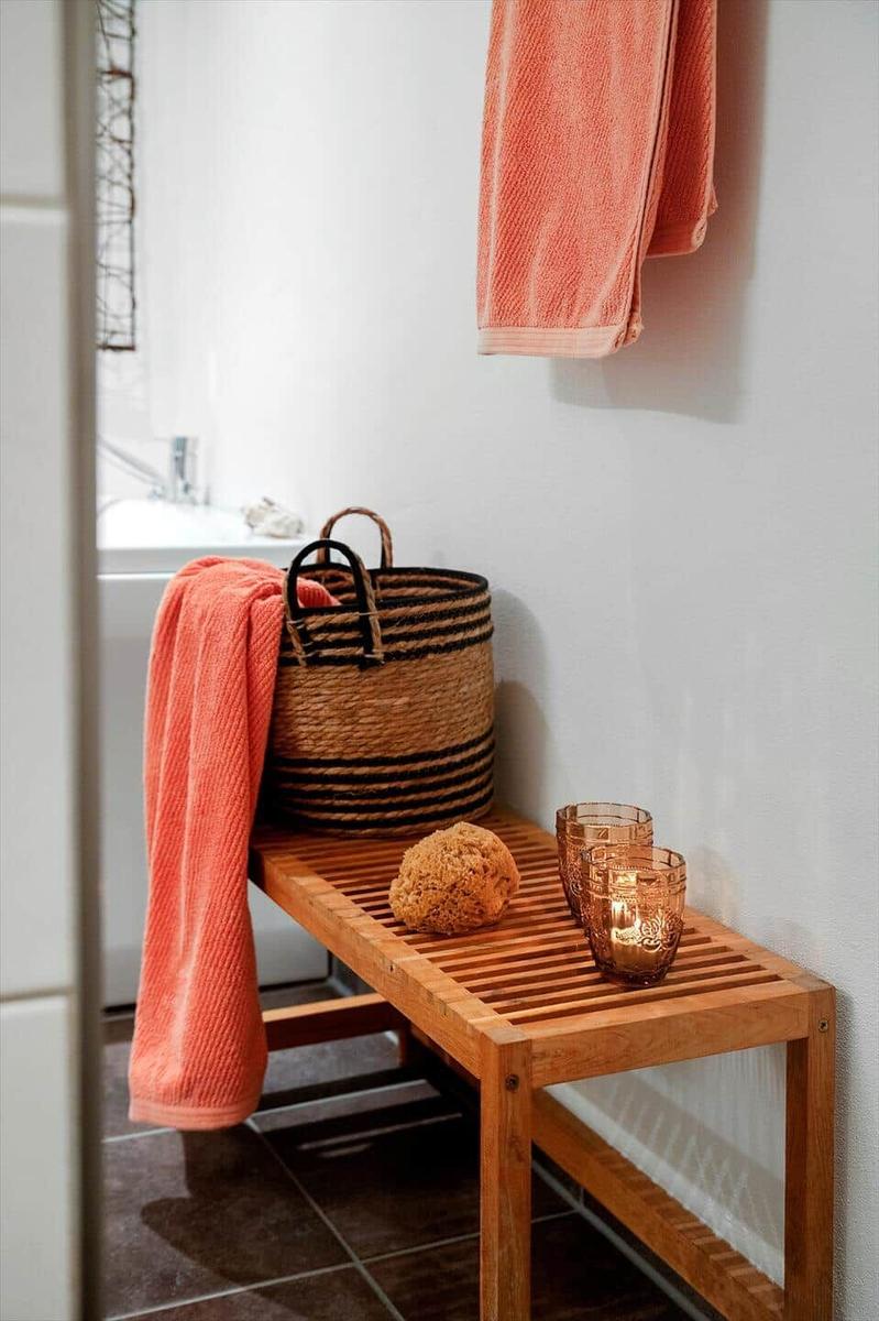 Holzbank mit Seifenschaum, Glas und Korb in Bad. Handtuch an Wand hängend.
