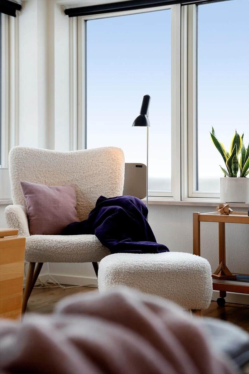Ein Sessel mit Kissen und Decke vor großen Fenstern mit Pflanze und Stehlampe.