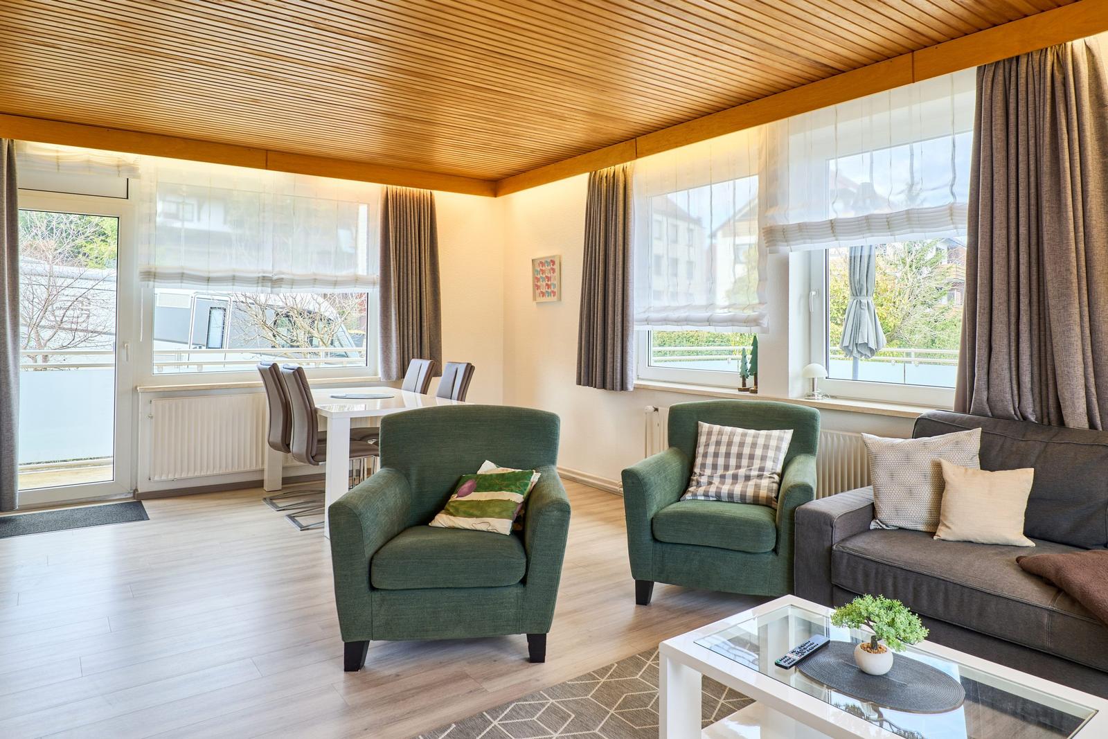 Wohn- und Essbereich mit grünen Sesseln, Sofa und Glas-Tisch. Holzdecke und große Fenster.
