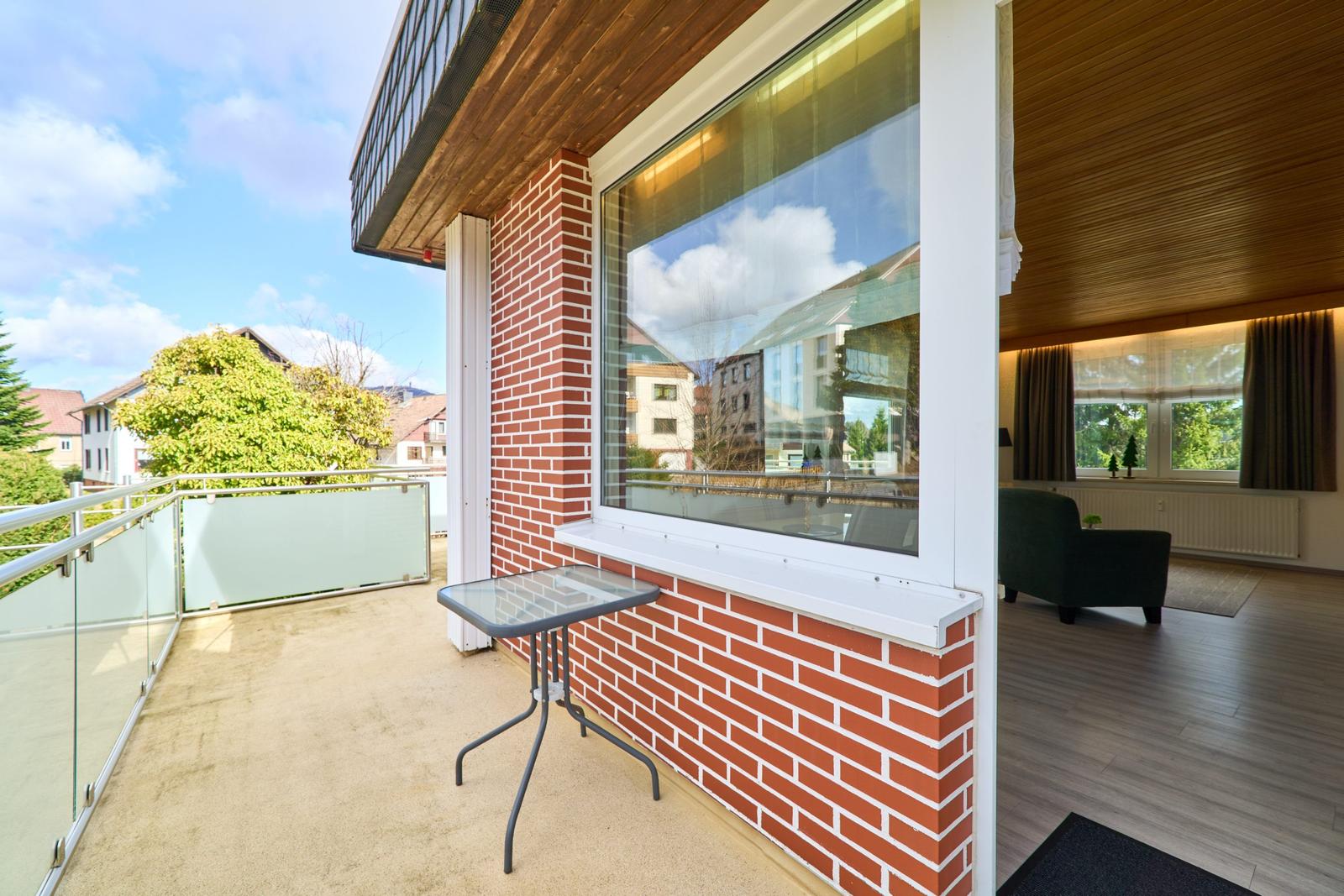 Terrasse mit Tisch und Blick auf Grünflächen. Wohnzimmer mit Sessel und Holzboden sichtbar.