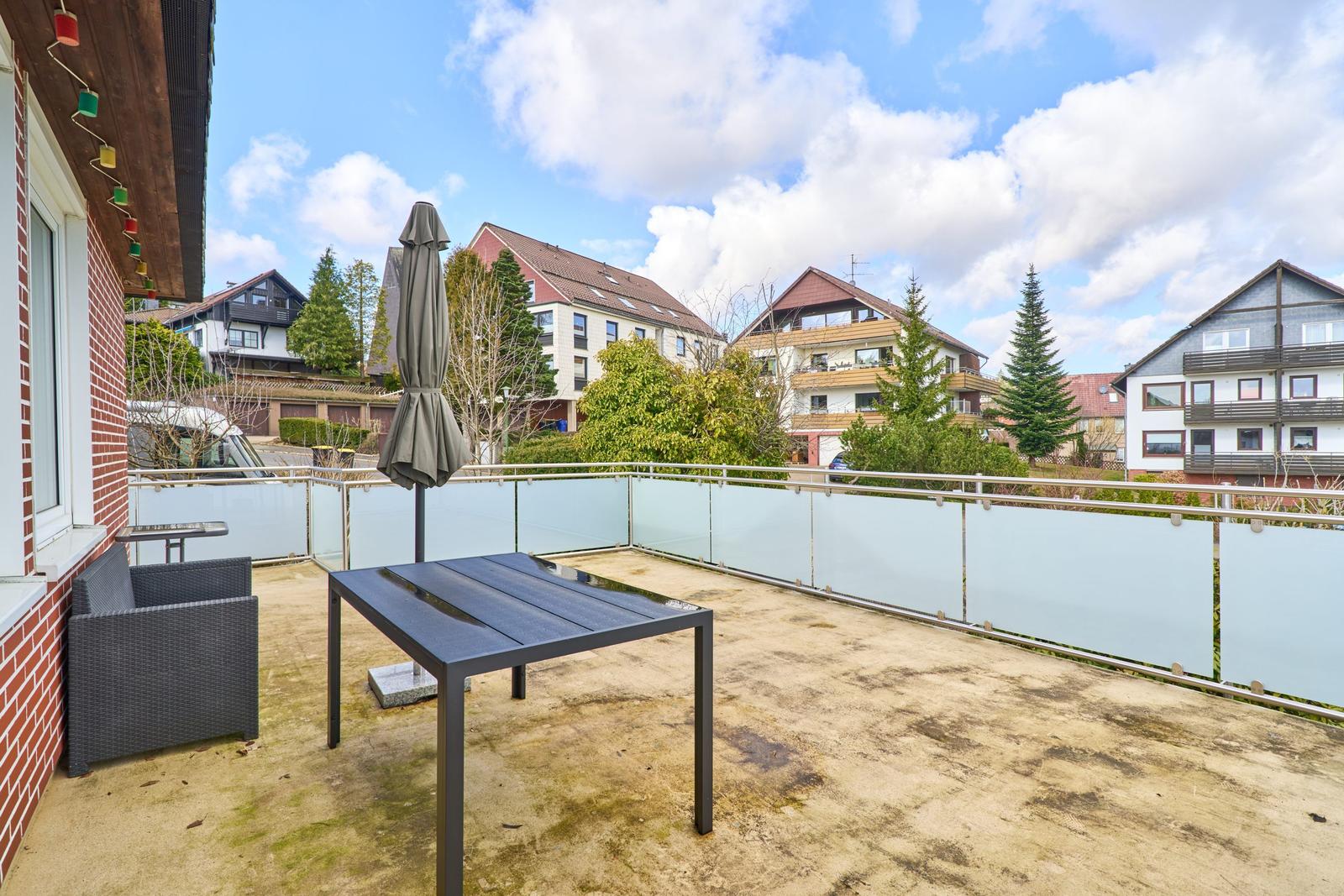 Terrasse mit Tisch, Stuhl und geschlossenem Sonnenschirm. Glasgeländer und Sicht auf Nachbarhäuser.