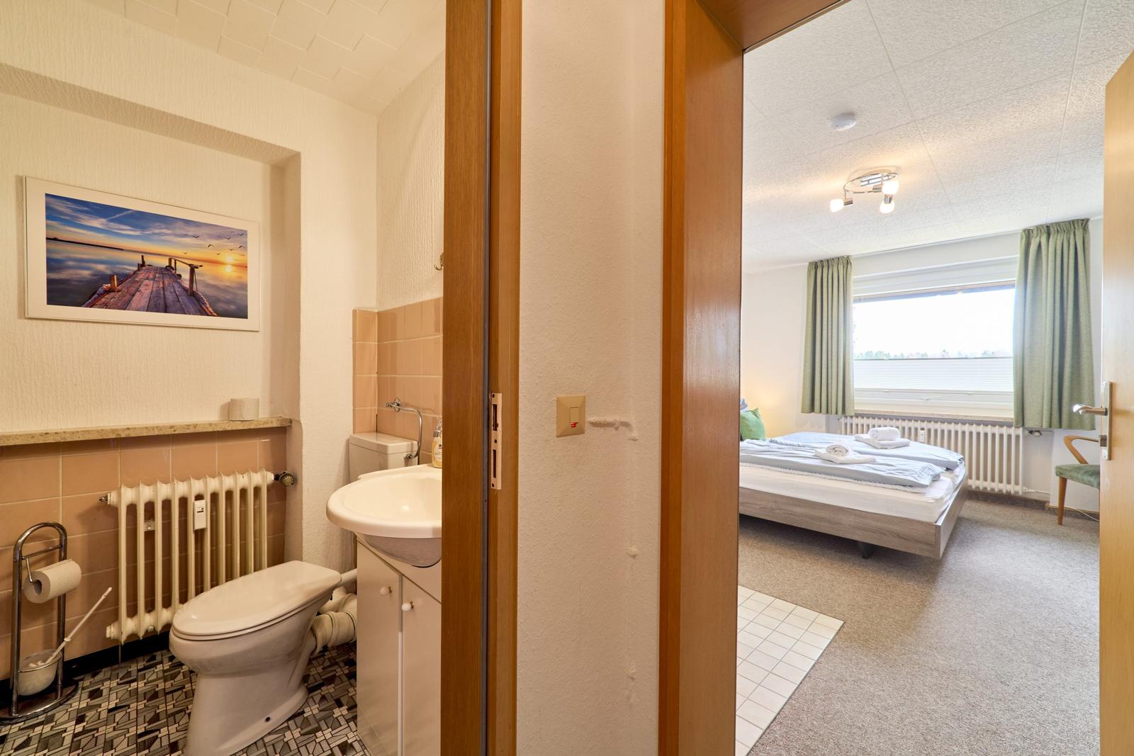 Badezimmer mit Toilette und Waschbecken. Blick in das Schlafzimmer mit Bett und Fenster.