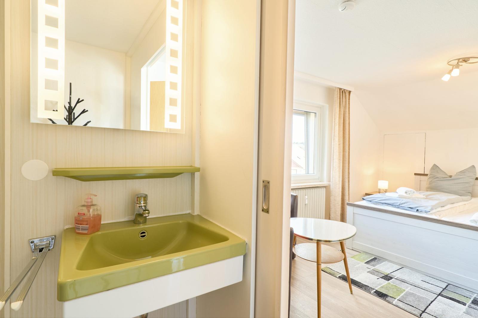 Badezimmer mit grünem Waschbecken und Spiegel. Blick in Schlafzimmer mit Bett und Tisch.