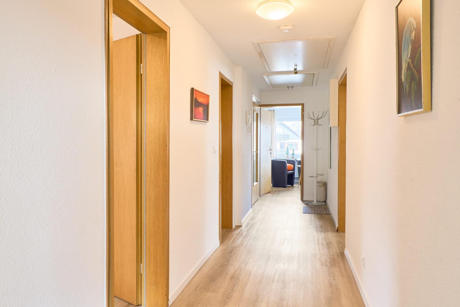 Flur mit Holzboden, Türen und Wandbildern. Durchgang zum Wohnbereich mit Sitzgelegenheit.