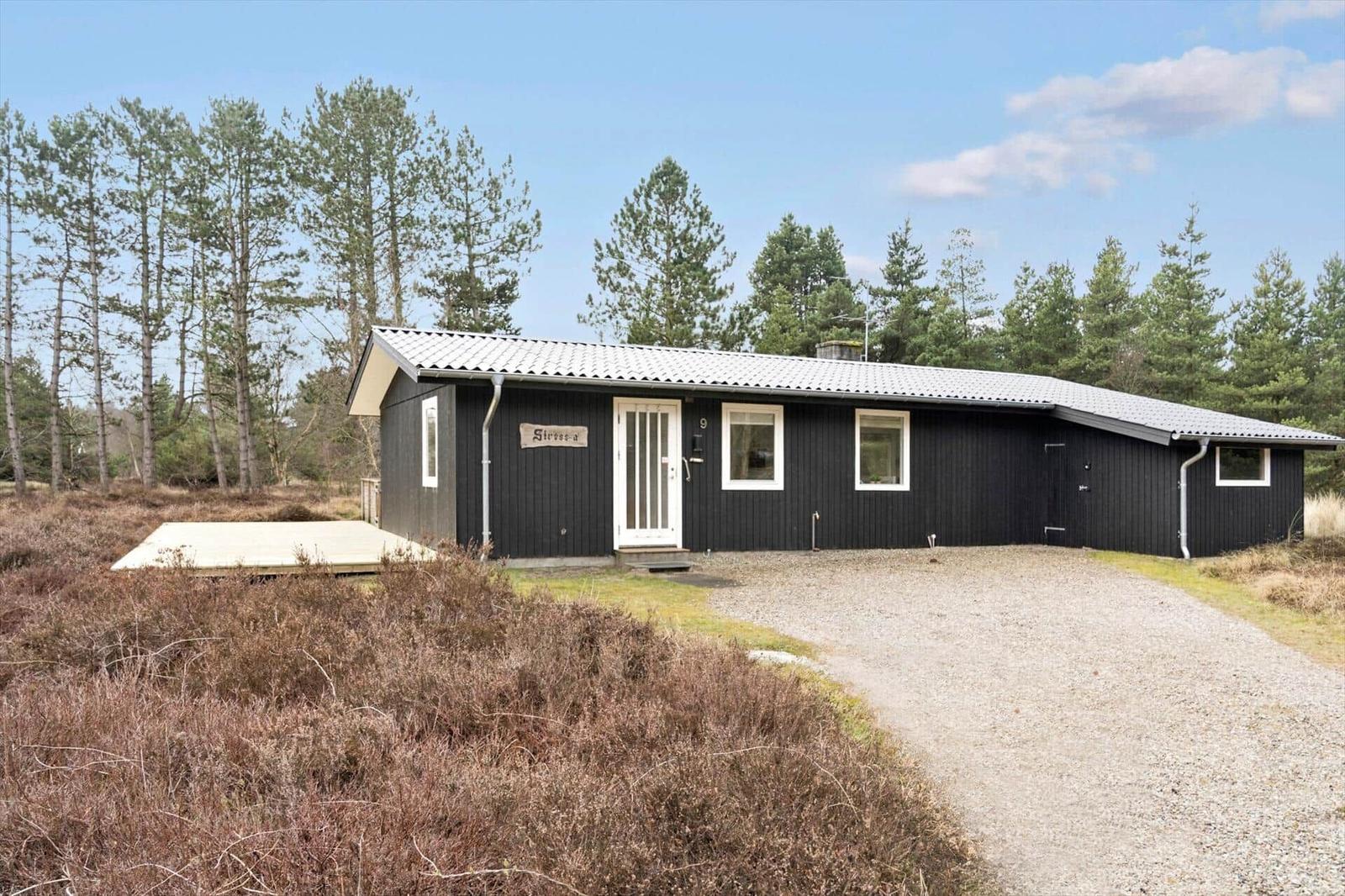 Schwarzes Haus mit weißer Tür und Fensterrahmen, umgeben von Bäumen und Heide.