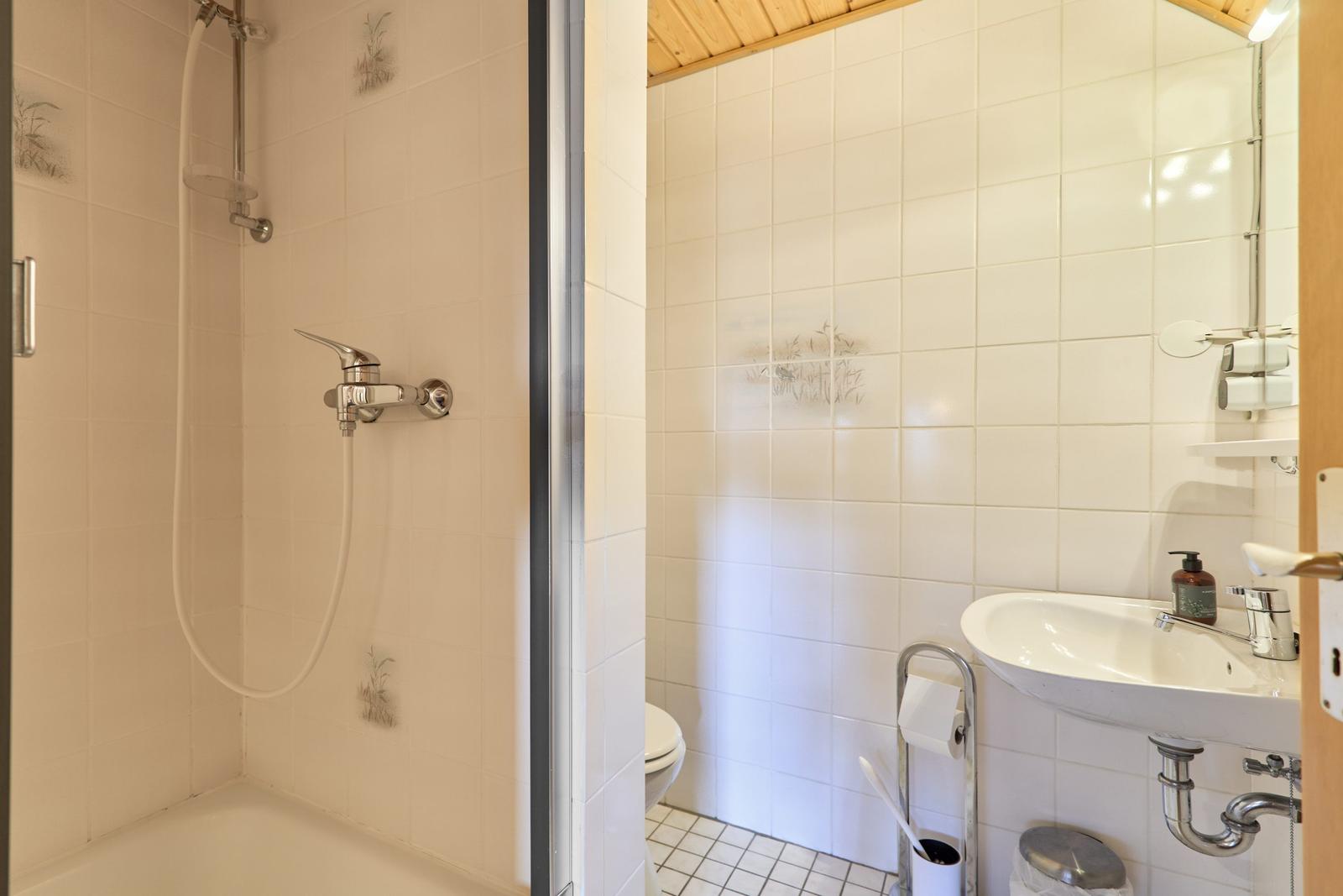 Badezimmer mit Dusche, Waschbecken und Toilette. Weiße Fliesen und Holzdecke.