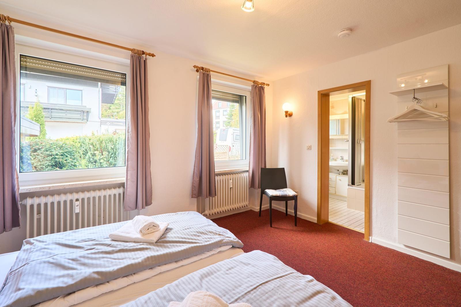 Zimmer mit zwei Betten, Fenstern und Blick auf Garten. Badezimmer und Kleiderständer sind sichtbar.