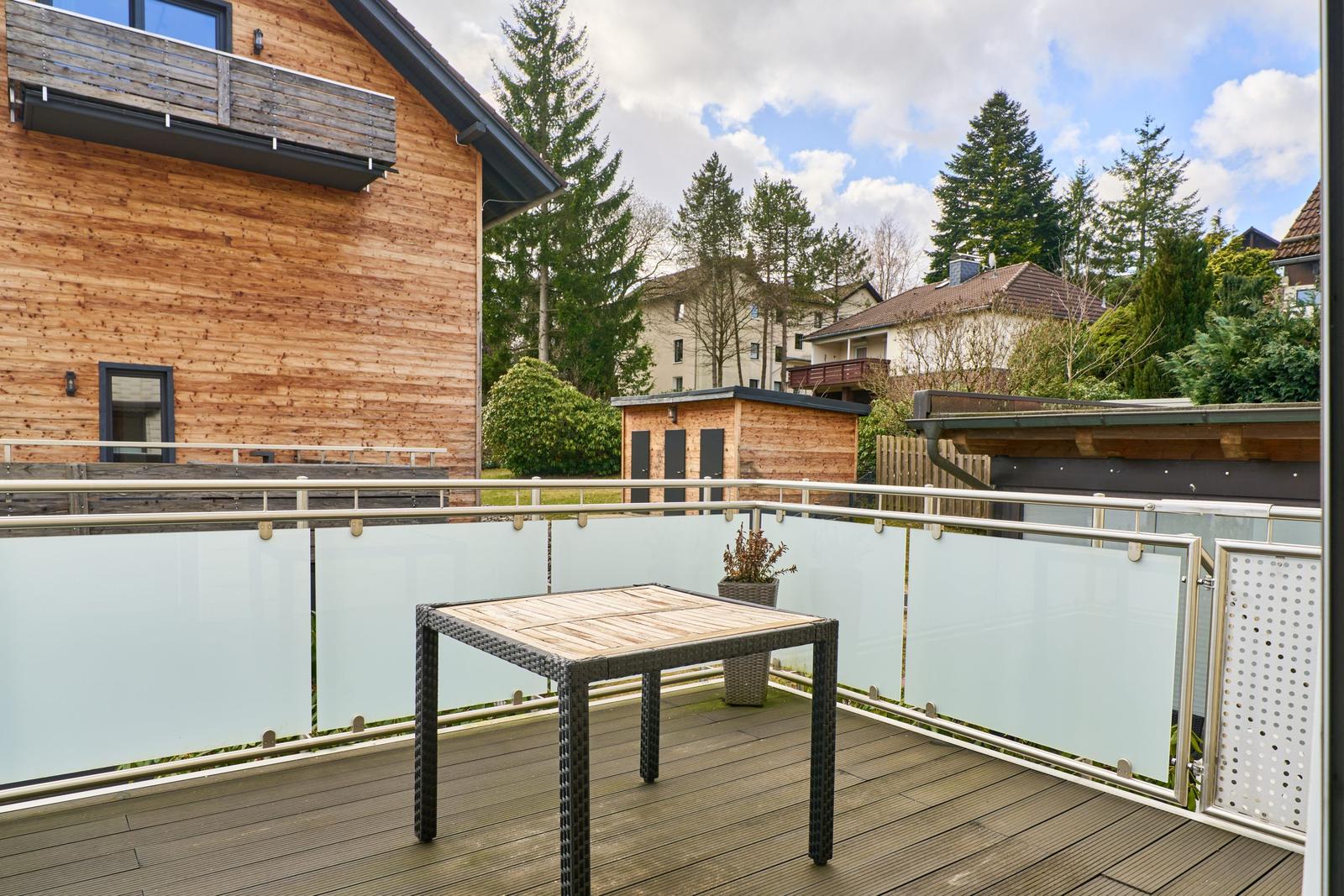 Terrasse mit Tisch, Glasgeländer und Blick auf Holzhaus und Bäume.