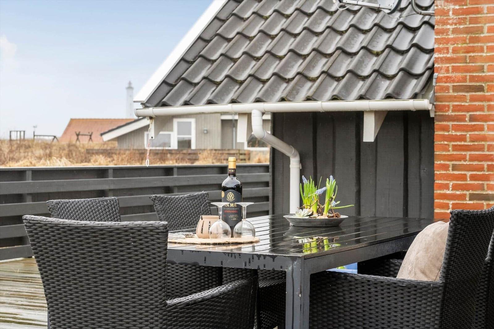 Terrasse mit Tisch und Stühlen, darauf Flasche und Blumen. Hintergrund: Haus mit Dach und Zaun.