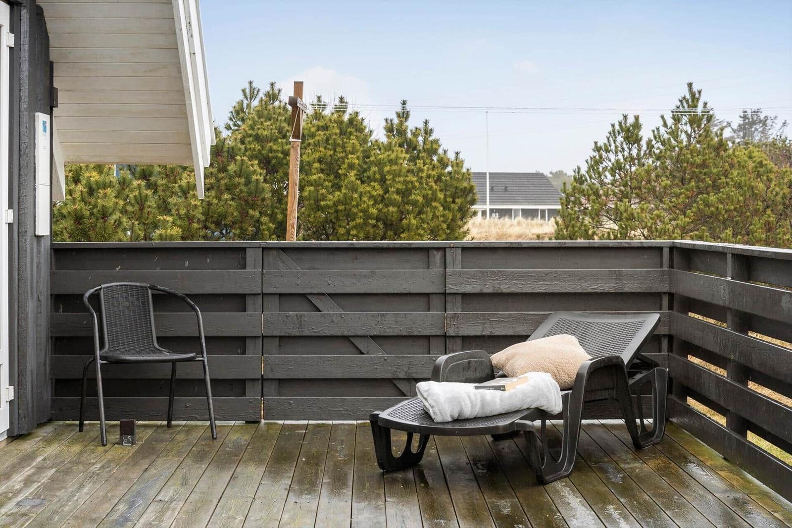 Terrasse mit Liegestuhl und Stuhl, umgeben von dunklem Holzgeländer und Bäumen.