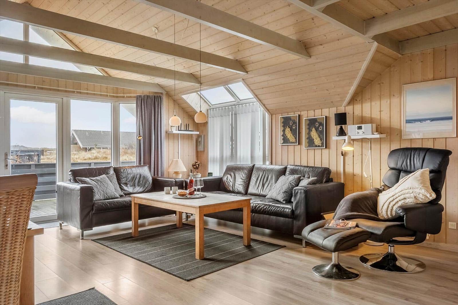 Wohnzimmer mit Ledersofa, Sessel und Holztisch. Holzwände und Dachbalken. Fenster mit Blick nach draußen.