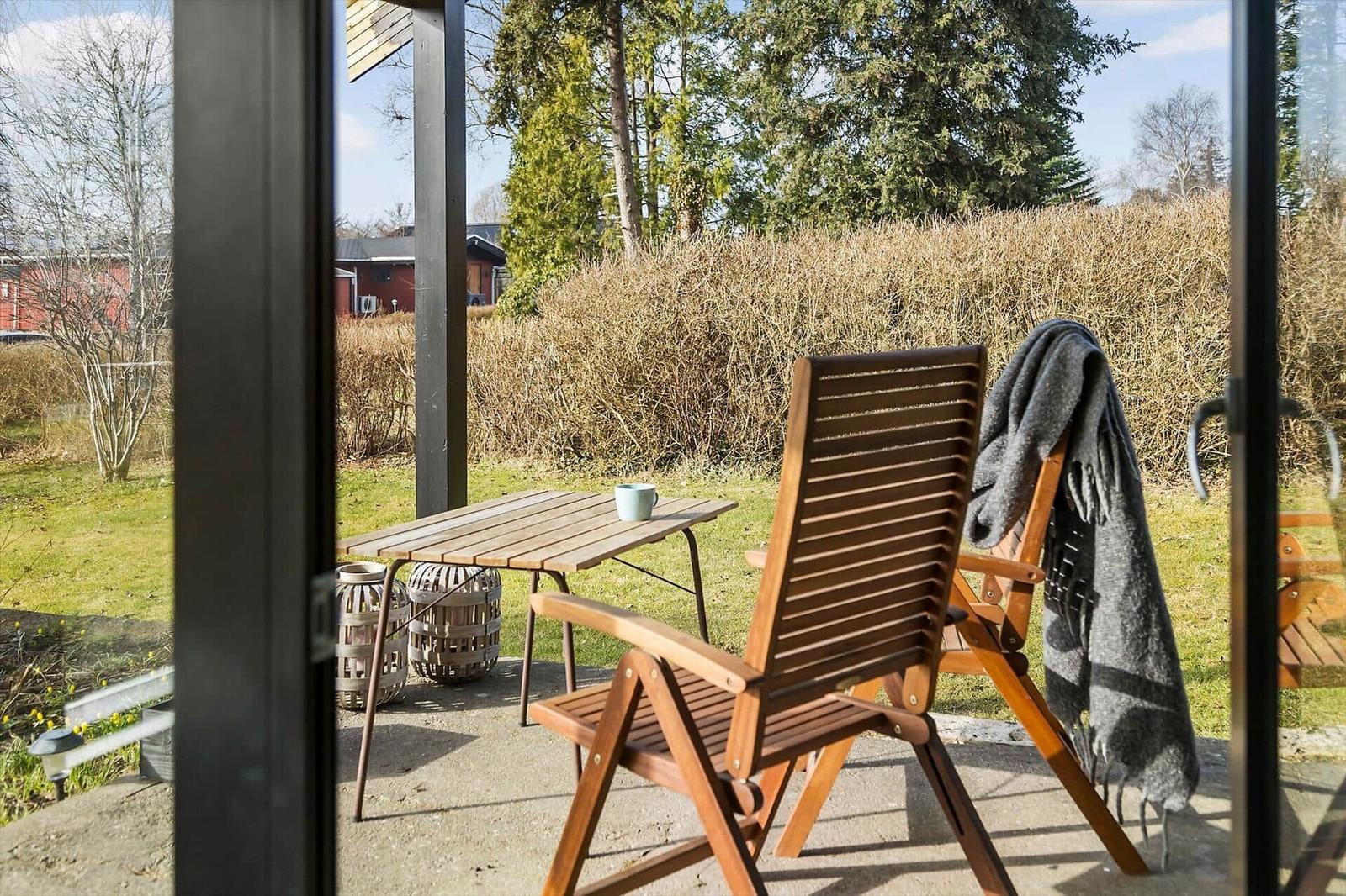 Terrasse mit Holzstuhl, Tisch und Decke. Hintergrund: Gras, Sträucher und Bäume.
