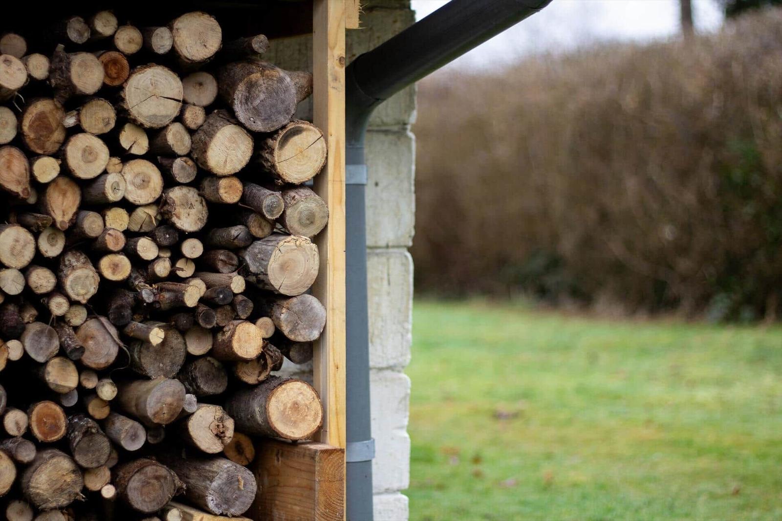 Holzstapel neben Steinmauer mit Gras und Sträuchern im Hintergrund.