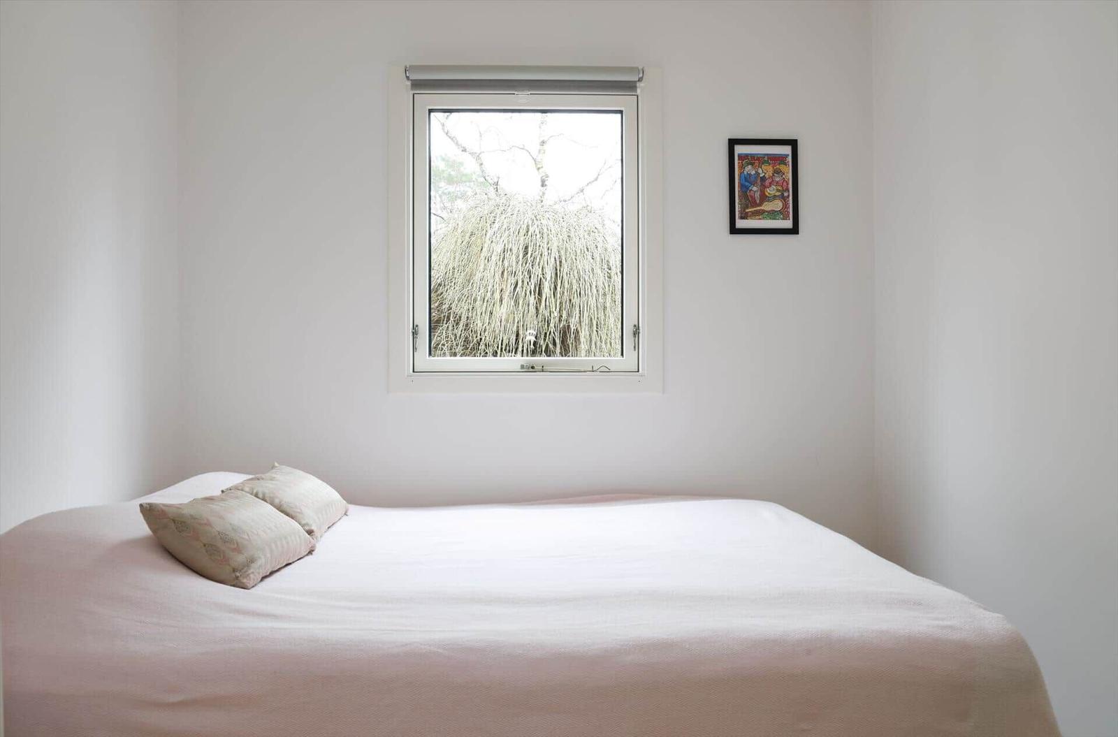 A bed with pillows under a window with a view of trees. A picture hangs on the wall.