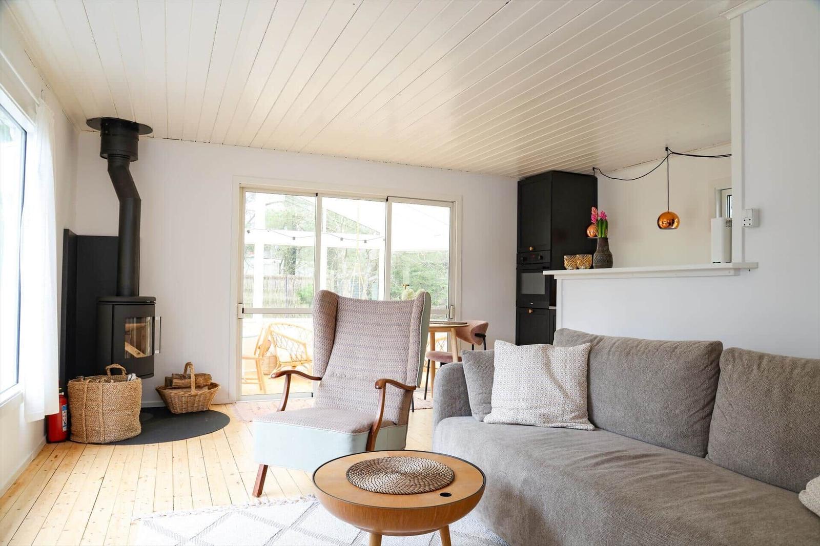 Living room with sofa, armchair, and wood stove. Wooden floor and large windows.