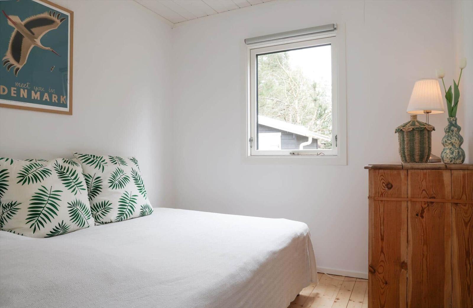 Bedroom with bed, window, and wooden dresser. Wall art and lamp are visible.