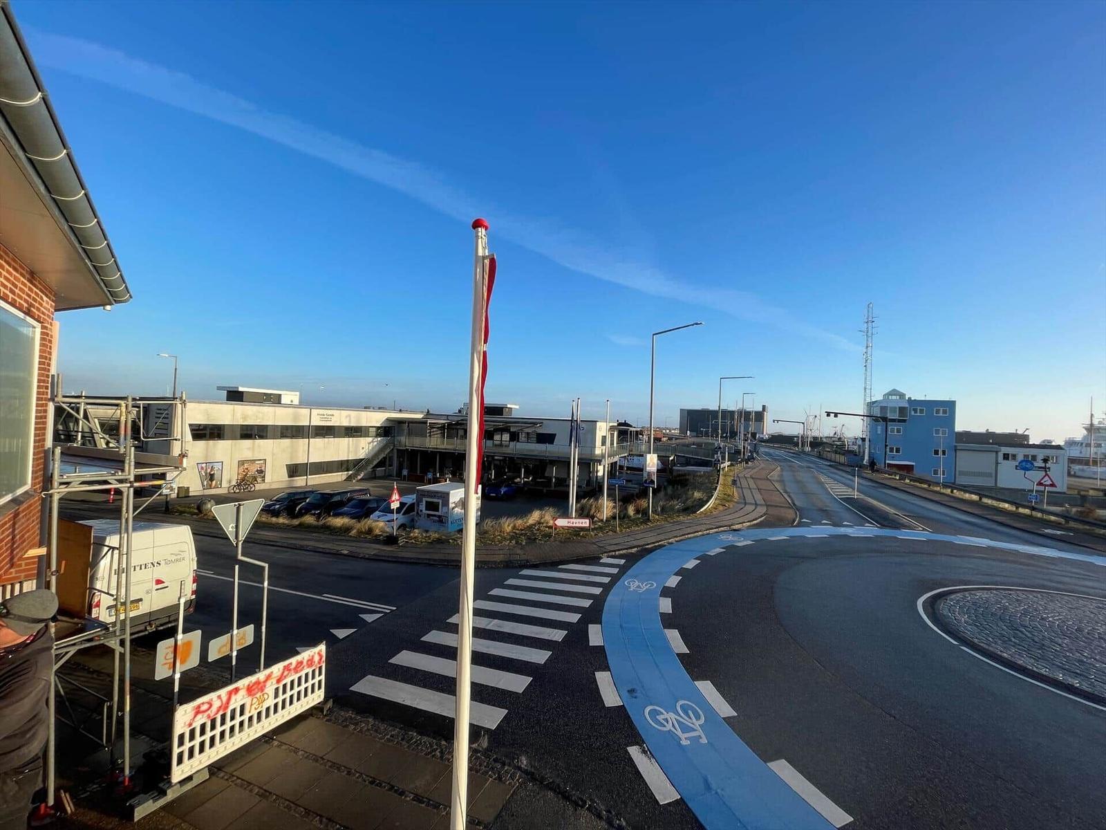 Ansicht von einer Straße mit Radweg und Flagge. Im Hintergrund sind Gebäude und ein Hafen zu sehen.