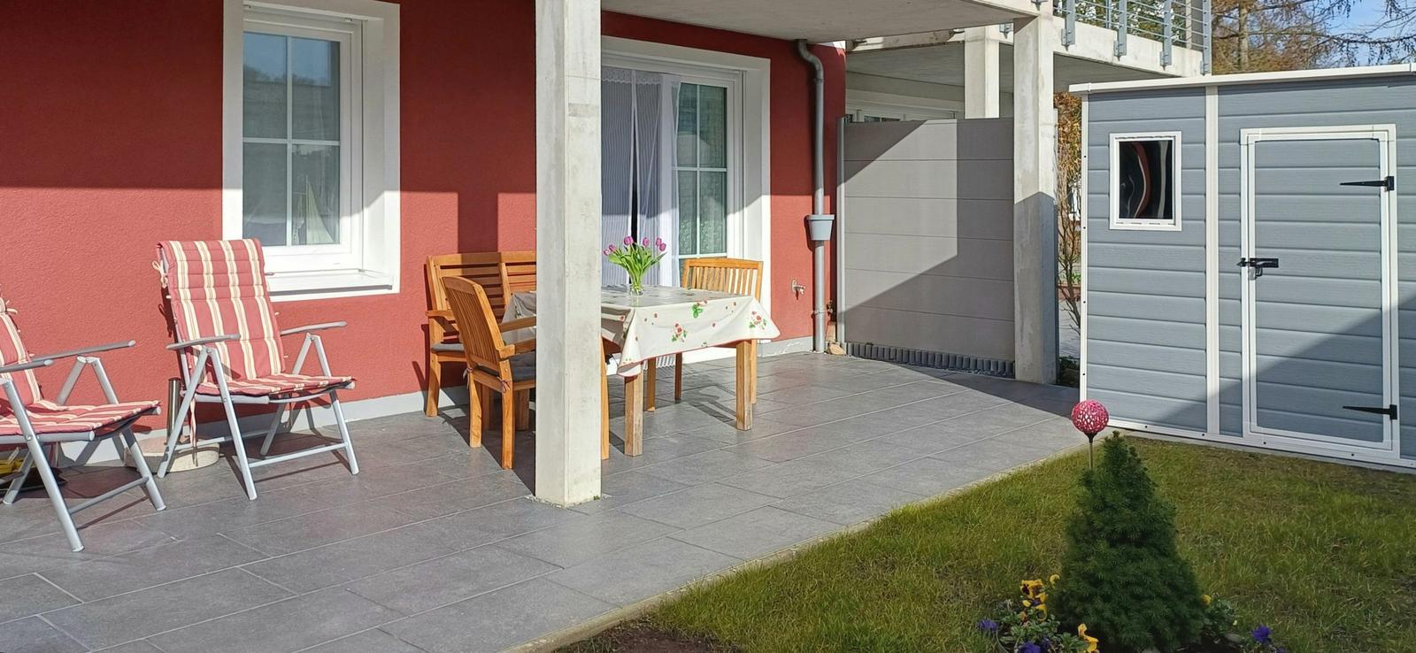 Terrasse mit Tisch, Stühlen und Sonnenschutz. Neben einem Gartenhaus mit Tür und Fenster.