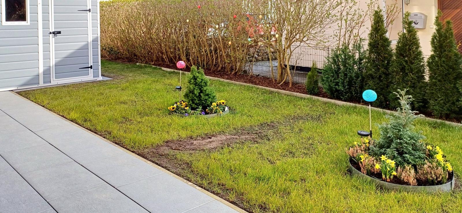 Garten mit Rasenfläche, zwei Blumenbeeten und einem Holzhaus.